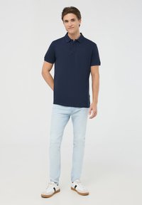 Granatowa koszulka polo z miękkiego materiału, z kołnierzykiem i dwiema guzikami, w połączeniu z jasnoniebieskimi dżinsami oraz białymi trampkami z czarnymi akcentami.