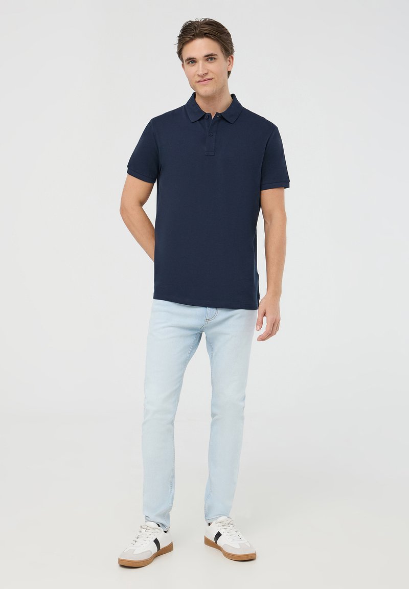 Granatowa koszulka polo z miękkiego materiału, z kołnierzykiem i dwiema guzikami, w połączeniu z jasnoniebieskimi dżinsami oraz białymi trampkami z czarnymi akcentami.