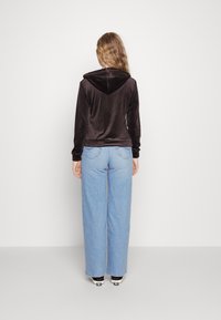 Hnědá velurová mikina s kapucí, kombinovaná s vysokými modrými širokými džíny a černými teniskami. Jednoduchý design bez viditelných vzorů.