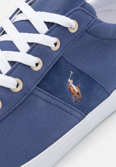 Tênis de lona azul-marinho com atacadores e ilhós brancos, apresentando um logo de jogador de polo bordado na lateral. Sola de borracha com uma textura suave.