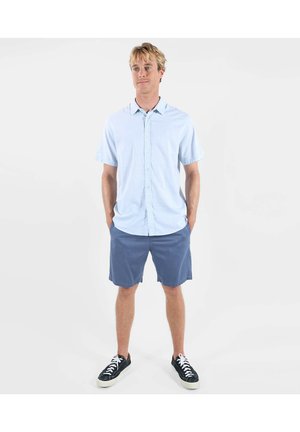 Jeune homme debout les mains dans les poches, portant une chemise à manches courtes bleu clair à boutons, un short bleu et des baskets noires sur fond blanc.