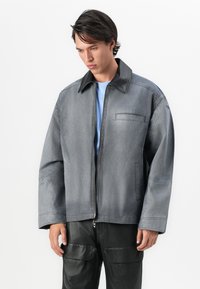 Νεαρός άνδρας με σκούρα μαλλιά, ντυμένος με μια φαρδιά γκρι ζακέτα με φερμουάρ και μαύρο γιακά, και μαύρα cargo παντελόνια, σε λευκό φόντο.