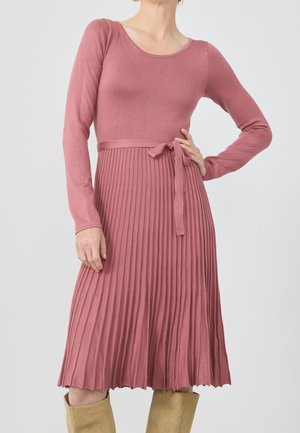 Kobieta ubrana w długą sukienkę z dzianiny w kolorze pudrowego różu z plisowanym dołem i przewiązanym paskiem, w połączeniu z beżowymi kozakami za kolano, stoi na gładkim tle.