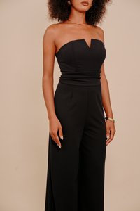 Kobieta w czarnym kombinezonie bez ramiączek z dekoltem w kształcie serca, nosząca złotą naszyjnik i bransoletkę, stojąca na beżowym tle.