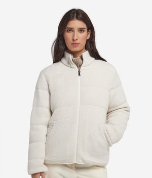 Femme aux longs cheveux foncés portant une veste blanche matelassée avec des poches devant et un col montant zippé, debout devant un fond blanc.