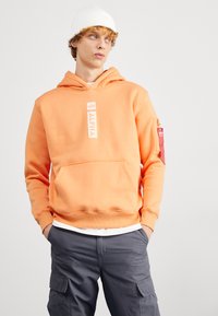 Sudadera de algodón naranja con bolsillo frontal, logo "ALPHA" y bolsillo lateral con cremallera. Combinada con pantalones cargo grises y un gorro blanco.