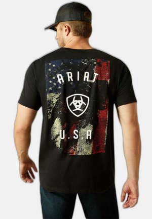 Homme portant une casquette noire et un T-shirt noir avec un drapeau américain usé, le texte "ARIAT" et un logo au dos, sur un fond uni.