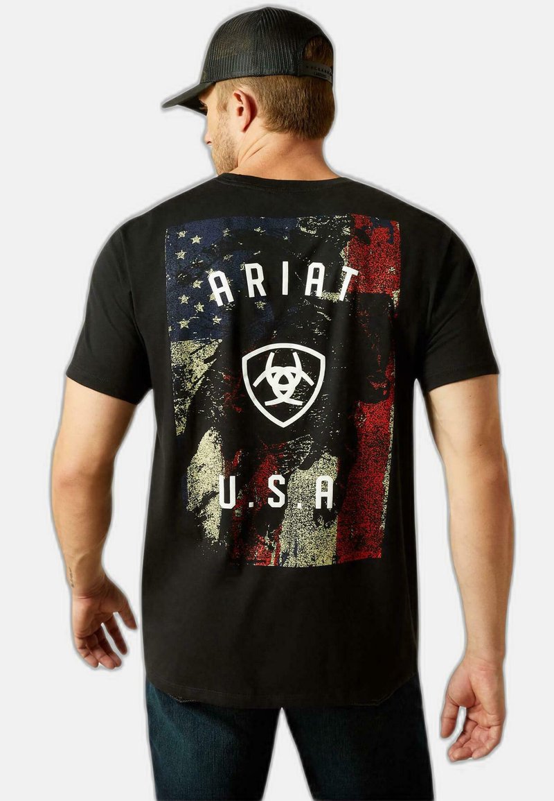 Hombre con gorra negra y camiseta negra con bandera americana desgastada, texto "ARIAT" y logo en la parte trasera sobre un fondo liso.