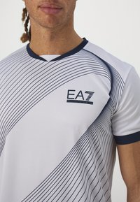 Bílé sportovní tričko s námořnickými akcenty, které má diagonální pruhovaný vzor, výstřih do V a logo EA7 na hrudi. Vyrobeno z hladké, elastické tkaniny.