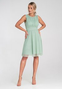 Jasnozielona sukienka bez rękawów wykonana z szyfonu, z dopasowanym gorsetem, plisowanymi detalami i rozkloszowaną spódnicą z przezroczystą nakładką.