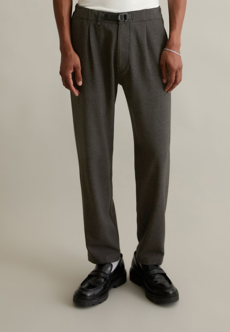 Gray pleated trousers with a belt, made of a textured fabric. Designed with front pockets and a tapered leg shape, paired with black shoes.