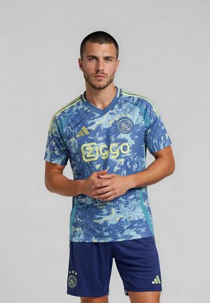 Hombre con equipación de fútbol del Ajax azul y amarilla de manga corta, de pie con las manos juntas frente a un fondo claro y liso.