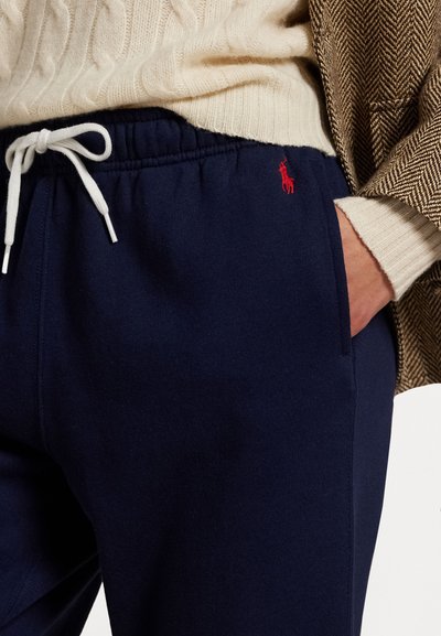 Navy fleece joggers with contrasting white drawstring. Features a small red logo on the left thigh and side pockets for functionality.