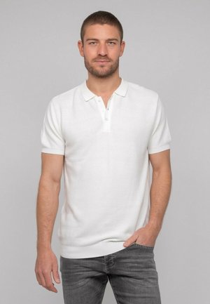 Homme aux cheveux bruns courts et à la barbe, portant un polo blanc à manches courtes et un jean gris foncé, debout devant un fond uni.