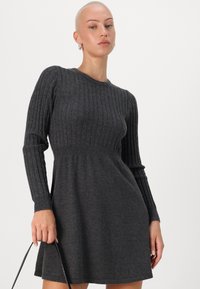 Tamsiai pilka raštuota megzta suknelė su ilgomisRankovėmis, prigludusia liemene, plisėta sijonu ir apvaliu iškirpimu. Modelis laiko juodą rankinę.
