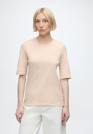 Blonde vrouw met bobkapsel draagt een lichtbeige top met korte mouwen en witte broek, staand tegen een effen witte achtergrond.
