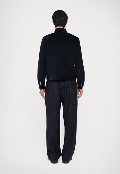 Veste en velours côtelé bleu marine avec col, poignets à boutons et détail à l'ourlet arrière, associée à un pantalon noir à rayures. Les deux vêtements sont ajustés.
