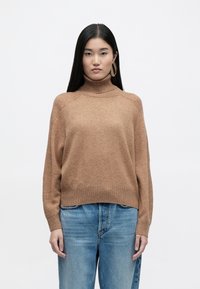 Καφέ πλεχτό πουλόβερ με ψηλό λαιμό και ριμπ κόψιμο στους καρπούς και το τελείωμα, συνδυασμένο με ψηλόμεσα μπλε τζιν. Λείο υλικό, χαλαρή εφαρμογή.