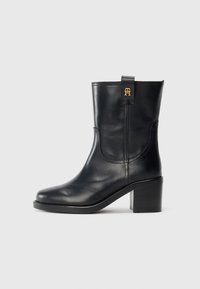 Botte en cuir noir mi-mollet avec talon bloc et logo "TH" doré sur la languette, sur fond blanc.