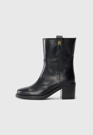 Botte en cuir noir mi-mollet avec talon bloc et logo "TH" doré sur la languette, sur fond blanc.