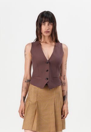 Brun vest uten ermer med V-hals, med en tettsittende design og tre svarte knapper, kombinert med et rutet skjørt i gult og brunt.