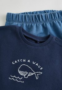 Pull en maille côtelée marine avec le texte blanc brodé "CATCH A WAVE" et un design de baleine, associé à un short bleu clair à taille élastique.
