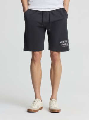 Uomo che indossa pantaloncini neri con coulisse e scritta bianca "Authentic Co." sulla gamba sinistra e sneakers bianche, in piedi con le mani nelle tasche.