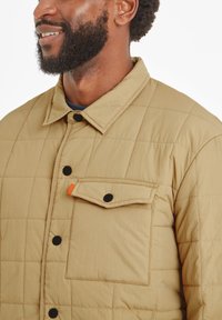 Quilted beige jacket with black snap buttons and a front pocket featuring an orange accent. Soft texture and structured design.