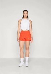 Valge lühike tops, oranžid lühikesed püksid ja valged tossud. Riietus sisaldab Nike logo, näidates sportlikke ja hingavaid materjale.