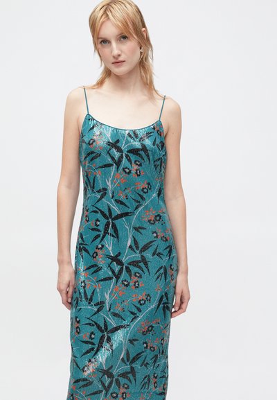 Mlada žena s plavom kosom nosi teal haljinu s šljokicama bez rukava, s tankim trakama i crnim i narančastim cvjetnim motivima, stoji nasuprot bijeloj pozadini.