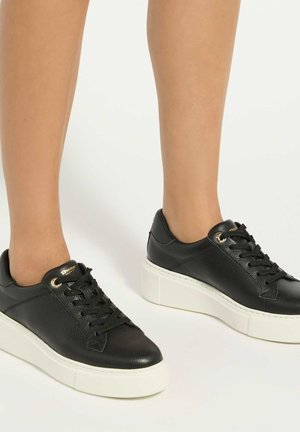 Baskets à plateforme en cuir noir avec semelles épaisses blanches portées sur des jambes nues sur fond blanc.