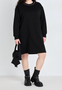 Černé šaty ve stylu sweatshirt s dlouhými rukávy a kulatým výstřihem, zdobené malými reflexními hřebíky. Kombinováno s černou taškou s mašlí a robustními černými botami.