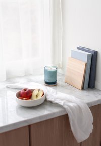 Blue glass candle on a marble countertop, accompanied by a white bowl of apples and two stacked books in blue and light wood.