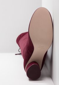 Bota de ante burdeos hasta la rodilla con punta redonda, tacón delgado y suela lisa. Presenta un detalle de cordones en el exterior a lo largo de la parte frontal.