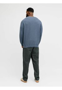 Blauer Strickpullover mit lockerem Schnitt, Rückennahtdetail; kombiniert mit dunkelgrünen Hosen und hellen Sandalen. Einfarbiger Hintergrund.