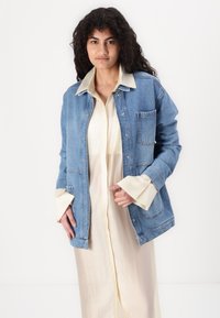 Lys blå denimjakke med avslappet passform, med en frontlomme, trykknappslukking og lagd med en kremfarget skjortekjole.