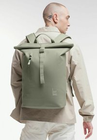 Plecak z rolowanym topem w stonowanej zielonej tkaninie, z kieszenią na zamek błyskawiczny, regulowanymi paskami i logo z boku. Gładka faktura, wodoodporny design.