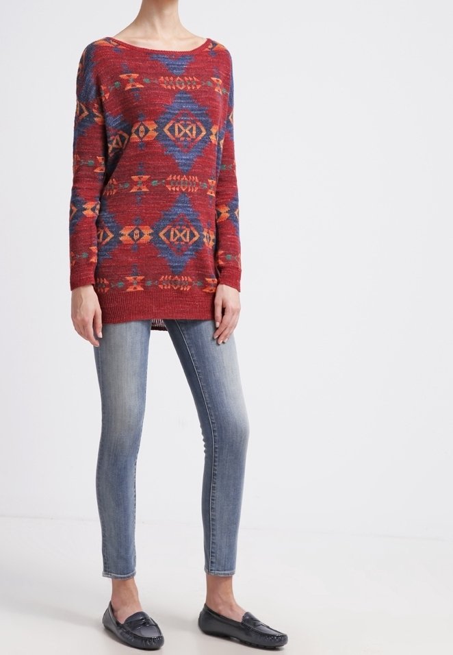 Roter Pullover mit Muster und blauen sowie orangenen geometrischen Designs, kombiniert mit hellblauen, eng anliegenden Jeans und dunklen Slippern.