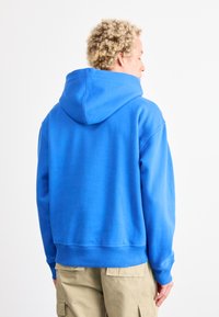 Sudadera con capucha azul, hecha de un tejido suave, con cuello alto, puños acanalados y un corte relajado. Mostrada desde la parte de atrás sin gráficos.