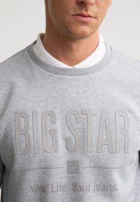Close-up af en person iført en grå sweatshirt med stor tekst "BIG STAR" og en lille stjerne-logo over sætningen "Your Life. Your Jeans."
