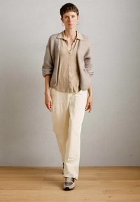 Donna che cammina in avanti indossando una camicia beige con bottoni, un cardigan color taupe chiaro, pantaloni crema e sneakers grigie su un pavimento di legno.