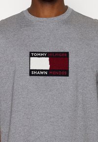 Harmaa puuvillainen crew-neck t-paita, jossa on suorakulmainen kirjottu patch, jossa on punaiset, valkoiset ja mustat bränditekstit Tommy Hilfigerille ja Shawn Mendesille.