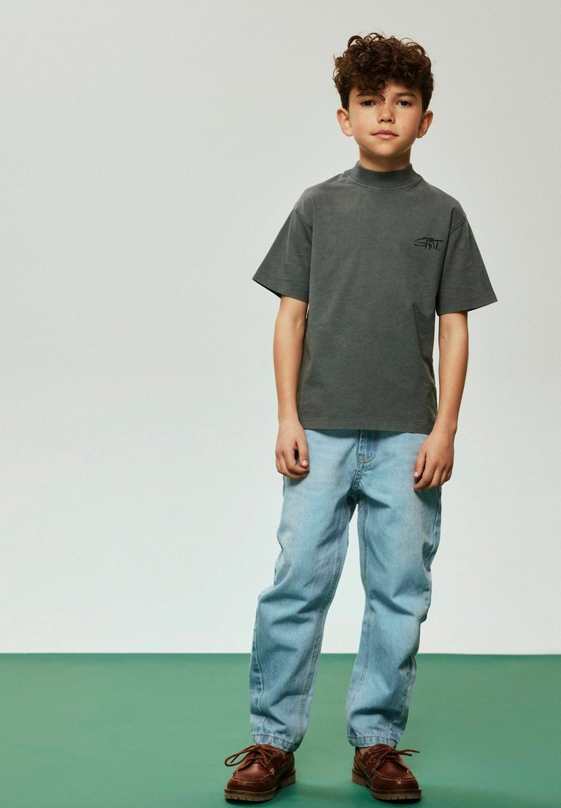 Gray short-sleeve t-shirt with high neckline and small black logo, paired with light blue denim jeans and brown lace-up shoes.