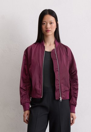 Vrouw met steil zwart haar draagt een donker bordeauxrode bomberjack met rits over een zwart topje en zwarte broek, staande tegen een lichte muur.