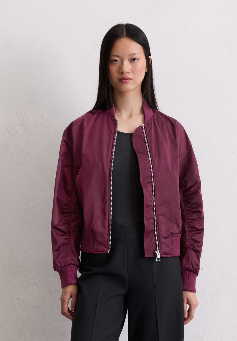 Woman with straight black hair wears a dark burgundy zip-up bomber jacket over a black top and black pants, standing against a light wall.