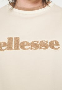 Beige sweatshirt med en texturerad beige logotyp som står "ellesse." Har en ribbad brun halsringning och en slät tygstruktur.