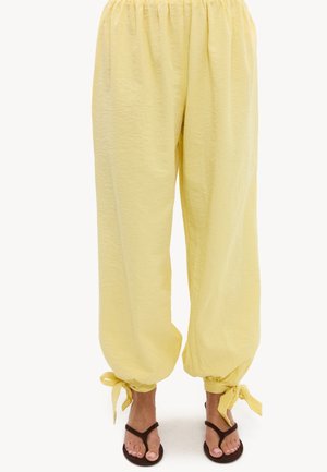 Pantalon ample jaune pâle avec taille élastique et poignets noués, porté avec des sandales tongs foncées sur un fond uni.