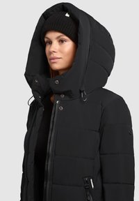 Black puffer jacket with a large, quilted hood. Features a front zipper, side pockets, and a smooth texture. Accented with metallic hardware.