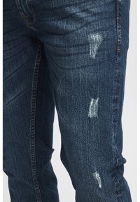 Jeans de mezclilla azul oscuro con detalles desgastados y parches deshilachados. Presentan un diseño clásico de cinco bolsillos y costuras visibles.