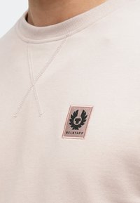 Lyserosa genser med ribbet hals, sømdetaljer og et svart emblemmerke med en vinget logo og teksten "BELSTAFF".